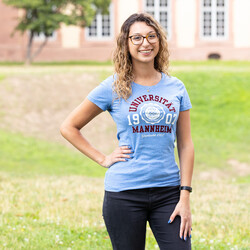 Eine lächelnde Person trägt ein hellblaues T-Shirt mit dem Logo der Uni Mannheim und steht auf der Mensawiese vor dem Schloss. Die Person heißt Anais Bentayeb.. 